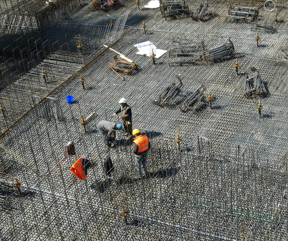 Widok z góry na plac budowy z grupą pracowników w kaskach i kamizelkach ochronnych, rozmawiających i przygotowujących zbrojenie betonu, symbolizujący pracę fizyczną na dużej inwestycji budowlanej.﻿