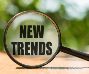 Szklana lupa powiększająca napis „NEW TRENDS” na drewnianym blacie na tle rozmytej zieleni, symbolizująca poszukiwanie nowych trendów.