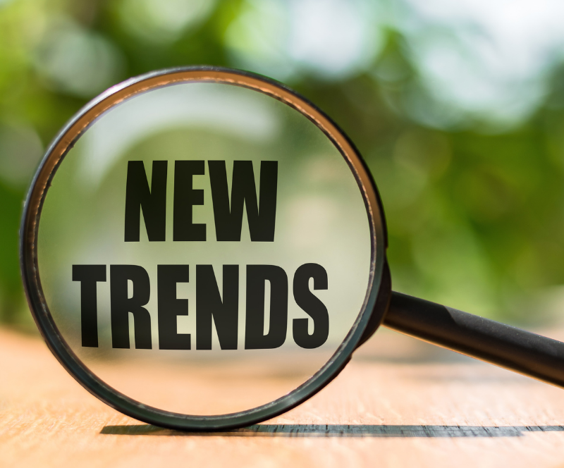 Szklana lupa powiększająca napis „NEW TRENDS” na drewnianym blacie na tle rozmytej zieleni, symbolizująca poszukiwanie nowych trendów.﻿